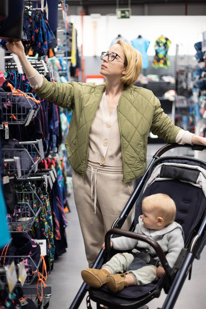 Casualy dressed mother choosing sporty shoes and clothes products in sports department of supermarket store with her infant baby boy child in stroller - Foto, Imagen