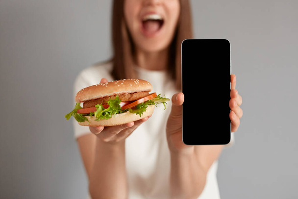 Image of unrecognizable unknown woman wearing white t-shirt holding fast food burger and showing mobile cell phone with blank screen isolated on gray background. - 写真・画像