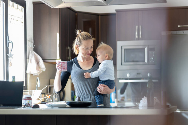 Ευτυχισμένη μητέρα να κρατάει το αγοράκι της πίνοντας πρωινό καφέ και φτιάχνοντας τηγανίτες για πρωινό στην κουζίνα. Οικογενειακός τρόπος ζωής, έννοια της οικογενειακής ζωής - Φωτογραφία, εικόνα