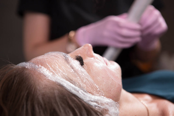 El esteticista en un salón de belleza envuelve la cara del paciente en celofán después de la anestesia. Procedimientos de cuidado y rejuvenecimiento de la piel. - Foto, imagen