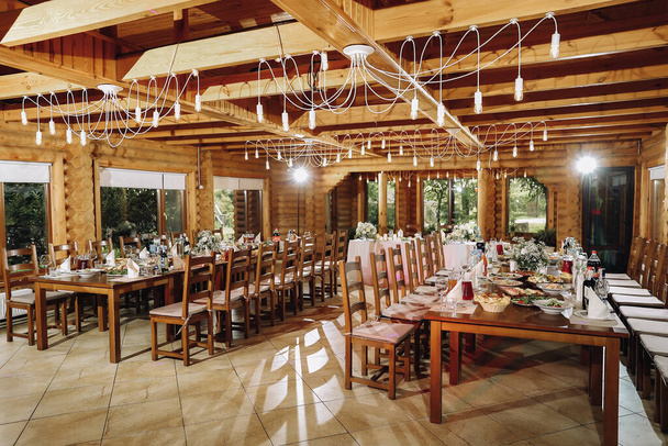 Banquet wooden hall for a rustic wedding with round decorated tables, Viennese chairs with flowered leaves and tablecloths. Horizontal. High quality photo - Фото, изображение