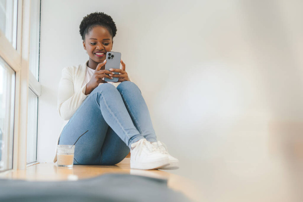 Gelukkig afro vrouw met ijs frisdrank zittend op cafe vensterbank met behulp van smartphone. Digitale technologie concept. Levensstijl van mensen. - Foto, afbeelding