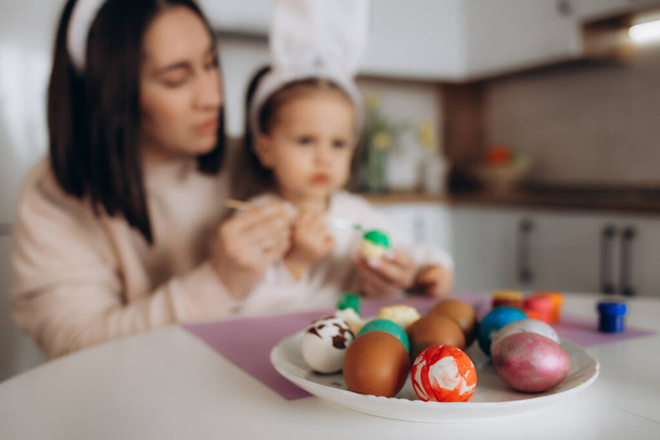 Щасливого Великодня. Мати та її дочка малюють великодні яйця. Щаслива сім'я готується до Великодня. Симпатична маленька дівчинка у вухах кролика на Великдень
. - Фото, зображення