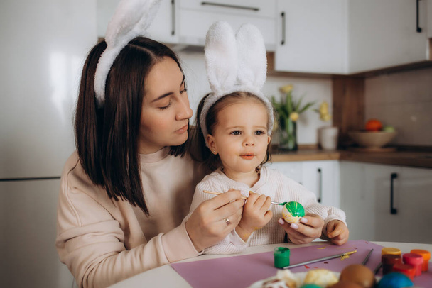 Szczęśliwego Wielkanocy. Matka i córka malują wielkanocne jajka. Szczęśliwa rodzina przygotowuje się do Wielkanocy. słodkie małe dziecko dziewczyna noszenie królicze uszy na Wielkanoc. - Zdjęcie, obraz