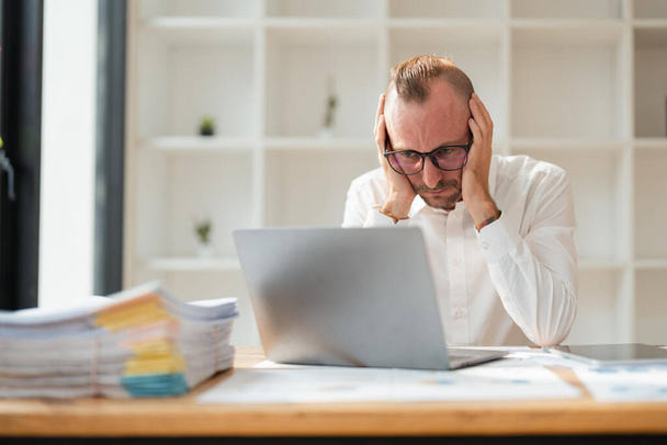 Κουρασμένος επιχειρηματίας κοιτάζοντας φορητό υπολογιστή, ενώ κάθεται στο γραφείο στο γραφείο. - Φωτογραφία, εικόνα