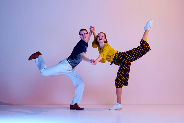 Happy emotional young couple, man and woman in stylish clothes dancing retro dance against gradient pink purple studio background. Concept of art, retro style, party, fun, movements, 60s, 70s culture - 写真・画像