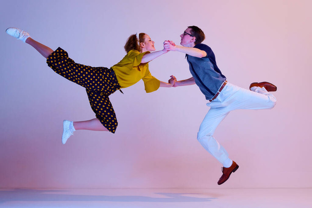 High jump. Young man and woman in stylish clothes expressively dancing retro dance against gradient pink purple studio background. Concept of art, retro style, party, fun, movements, 60s, 70s culture - Фото, изображение