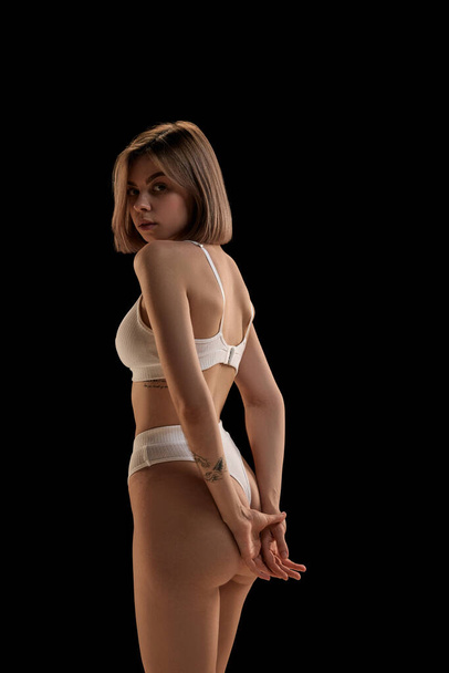 Young slim woman wearing white cotton lingerie posing over dark background. Concept of natural beauty, body and skin care, health, spa, cosmetics. Copy space for ad - Fotoğraf, Görsel