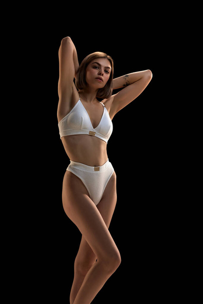 Young slim sensual woman wearing white inner wear posing over dark background. Concept of dieting, active and healthy lifestyle, natural beauty, body and skin care. Copy space for ad - Foto, Imagem