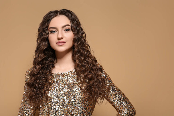 Beautiful young woman with long curly brown hair in golden sequin dress on beige background - Fotografie, Obrázek