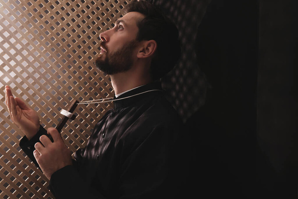 Catholic priest in cassock holding cross and praying to God in confessional - 写真・画像