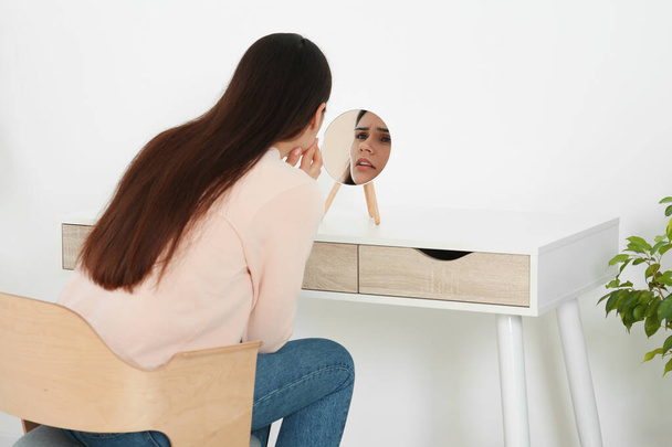 Young woman looking in mirror and touching her face indoors. Hormonal disorders - Foto, imagen