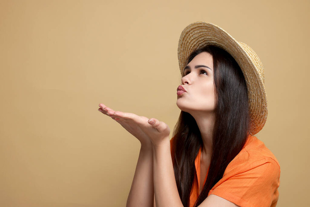 Красива молода жінка в солом'яному капелюсі дме поцілунок на бежевому тлі. Простір для тексту
 - Фото, зображення