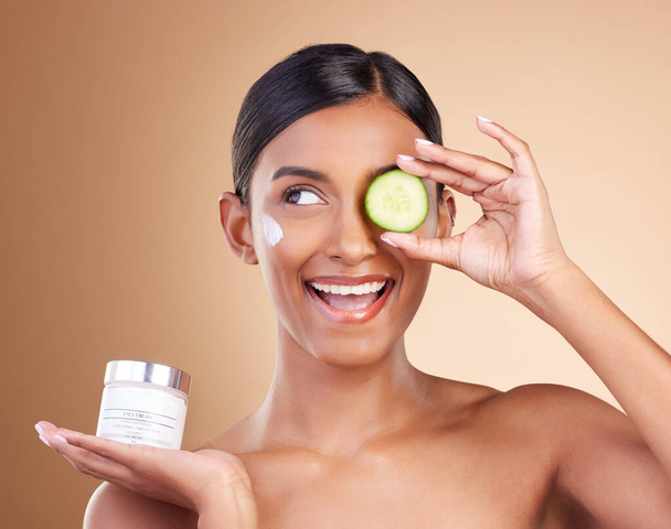 Beauty, skincare and Indian woman with cucumber, cream and dermatology against a brown studio background. Female, cosmetics and happy lady with natural care, lotion and healthy detox with grooming. - Фото, изображение