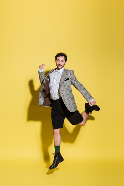 Trendy and smiling model in shorts and jacket jumping on yellow background  - Zdjęcie, obraz