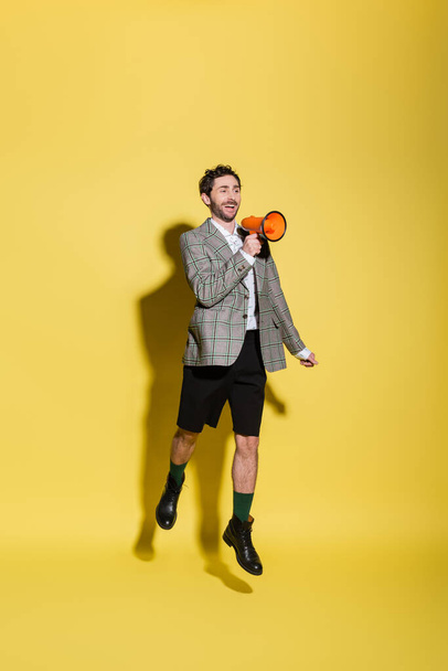 Cheerful and stylish man in shorts and jacket holding loudspeaker while jumping on yellow background  - Fotoğraf, Görsel