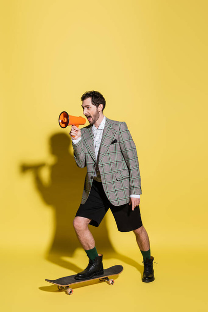 Trendy man holding loudspeaker and skateboarding on yellow background  - Valokuva, kuva