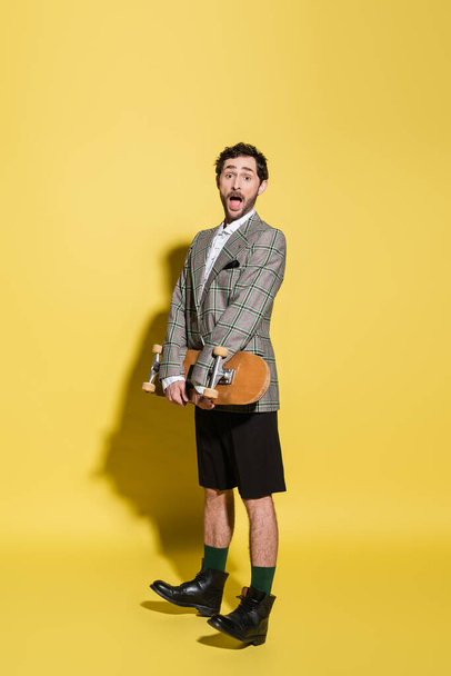 Excited and stylish man in jacket and shorts holding skateboard on yellow background  - Фото, зображення