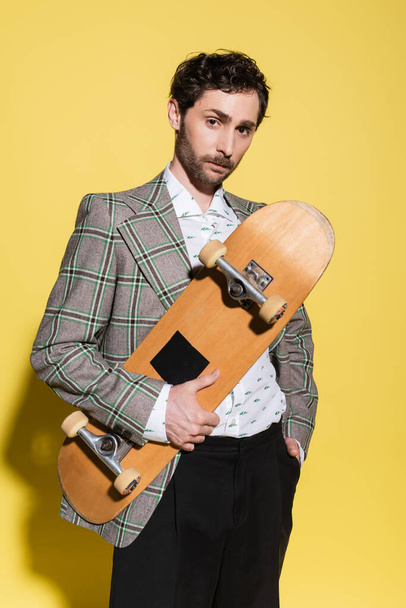 Trendy man in checkered jacket holding skateboard and posing on yellow background  - Foto, immagini