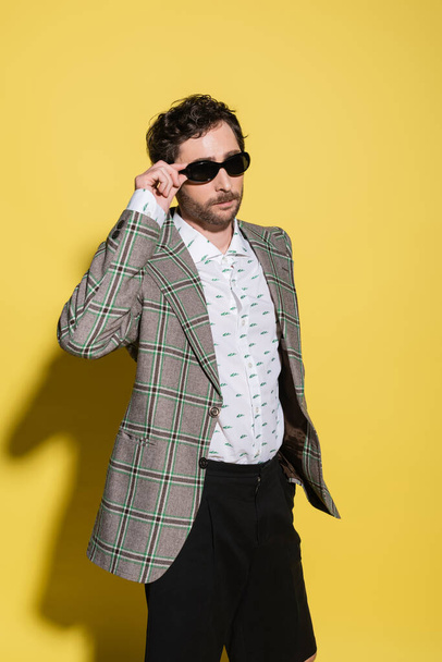 Trendy charming guy in jacket holding sunglasses on yellow background  - Фото, изображение