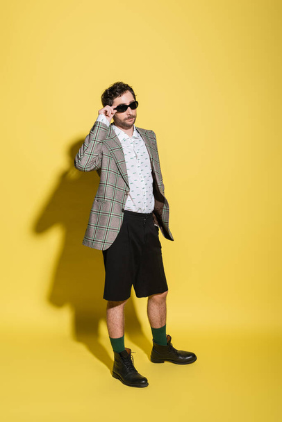 Fashionable man in shorts and jacket holding sunglasses on yellow background  - Fotografie, Obrázek