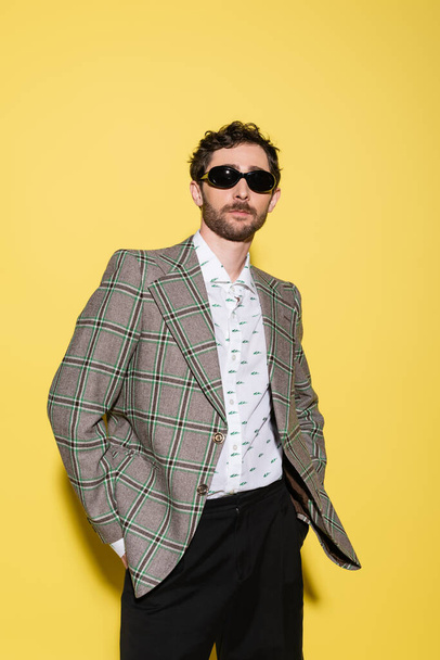 Trendy model in sunglasses and plaid jacket standing on yellow background  - 写真・画像