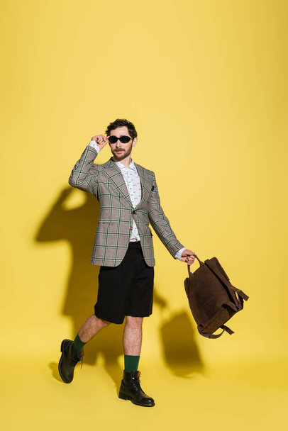 Fashionable model in sunglasses holding backpack and walking on yellow background  - Fotografie, Obrázek