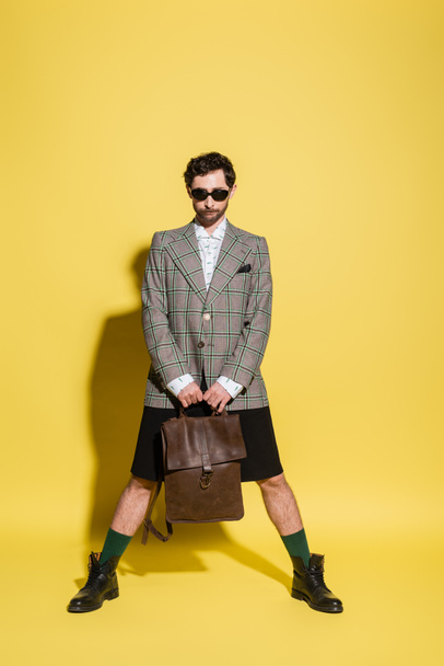Fashionable model in jacket and sunglasses holding bag while standing on yellow background  - Photo, Image