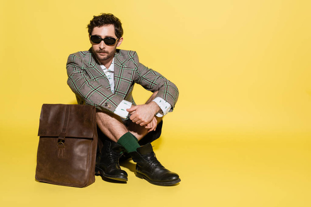 Trendy model in jacket and sunglasses sitting near bag on yellow background  - Photo, Image