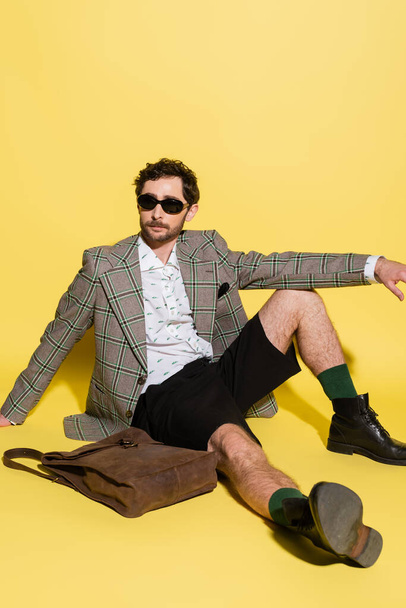 Trendy model in sunglasses and shorts sitting near bag on yellow background  - Foto, Imagem