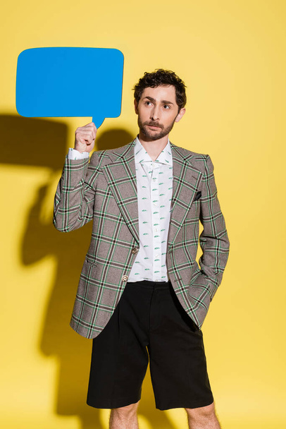 Trendy man in jacket and shorts holding speech bubble on yellow background  - Foto, Imagen