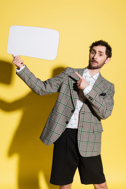 Charming guy in jacket pointing at empty speech bubble on yellow background  - Fotografie, Obrázek