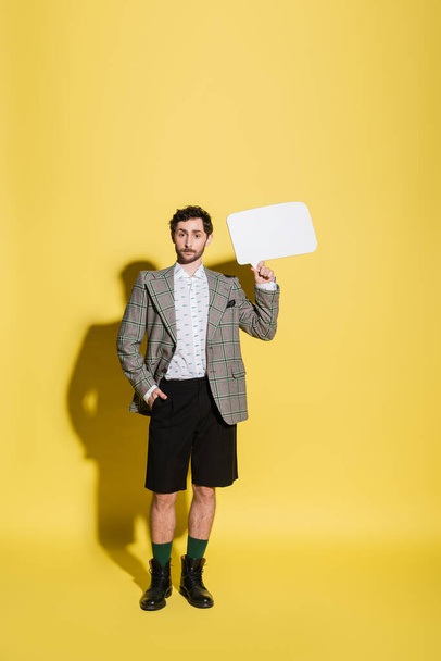 Full length of trendy model in shorts and jacket holding speech bubble on yellow background  - Fotografie, Obrázek