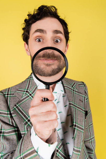 Wide angle view of stylish man in jacket holding magnifying glass isolated on yellow   - Photo, Image