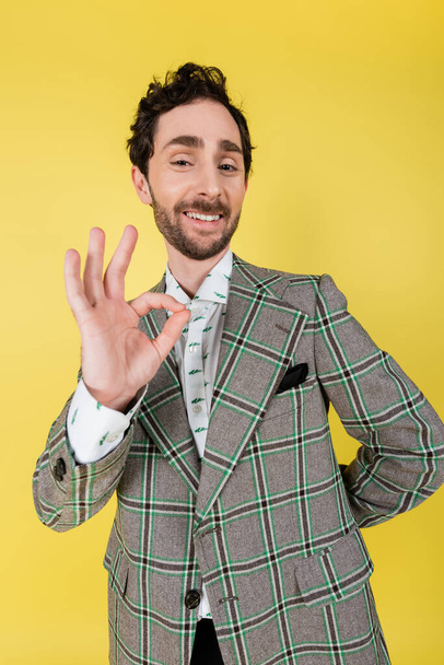 Wide angle view of smiling and stylish man showing ok gesture isolated on yellow   - Photo, image