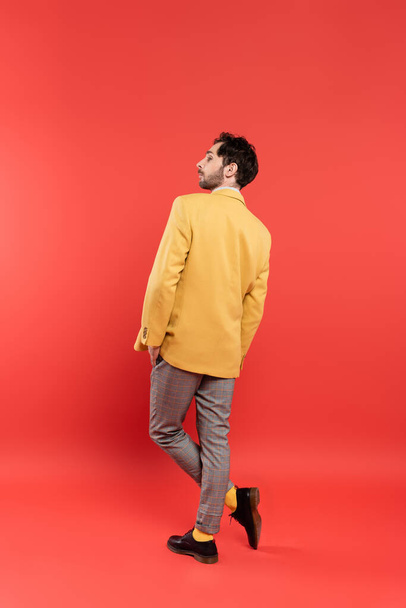 Πλευρική άποψη του κομψού άνδρα σε κίτρινο σακάκι θέτει σε κοραλλί κόκκινο φόντο - Φωτογραφία, εικόνα