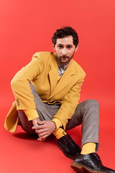 Trendy and brunette man in yellow jacket sitting on coral red background - Fotografie, Obrázek