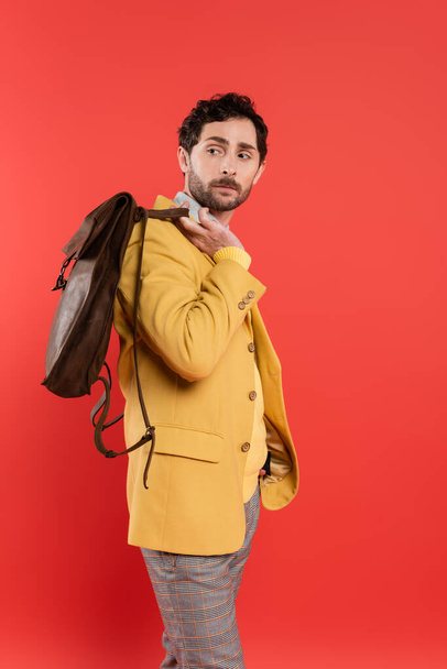 Trendy man in jacket holding brown backpack isolated on coral red  - Photo, Image