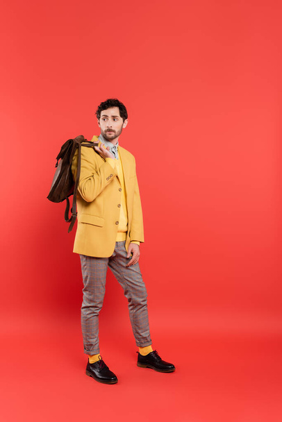 Full length of stylish model in yellow blazer holding backpack on coral red background - Φωτογραφία, εικόνα