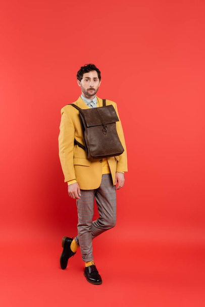 Full length of fashionable model with brown backpack over jacket looking at camera on coral red background - Valokuva, kuva
