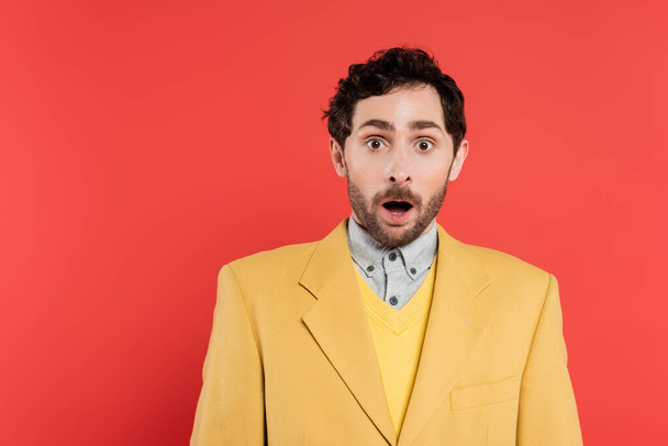 shocked man in yellow blazer standing with opened mouth and looking at camera on coral background  - Fotoğraf, Görsel