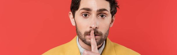 man in yellow blazer holding finger near lips while showing hush sign isolated on red coral background, banner  - Photo, Image
