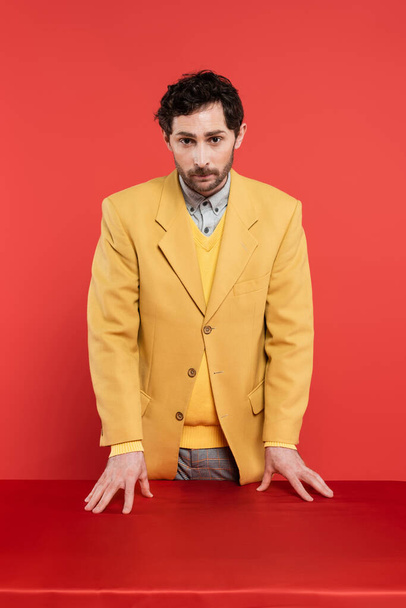 άνδρας σε κομψό κίτρινο σακάκι στέκεται κοντά στο κόκκινο γραφείο και κοιτάζοντας κάμερα σε κοράλλι φόντο  - Φωτογραφία, εικόνα