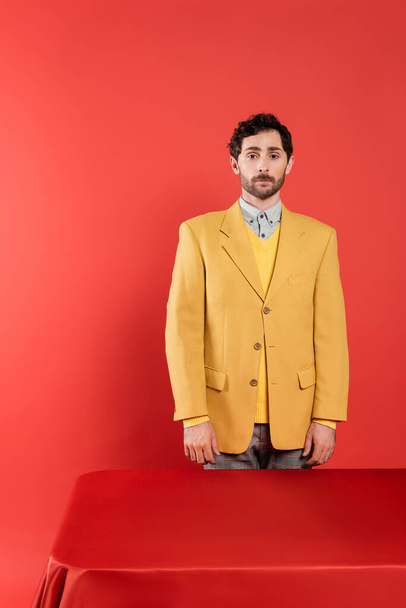 curly man in stylish yellow blazer standing near red desk and looking at camera on coral background  - Фото, изображение
