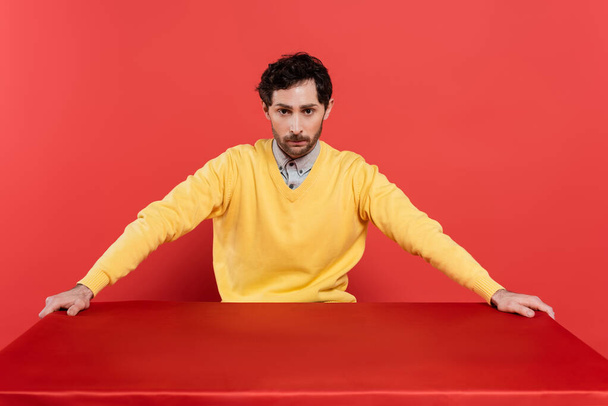 charming guy in yellow long sleeve jumper sitting at table with red tablecloth on coral background  - Fotografie, Obrázek
