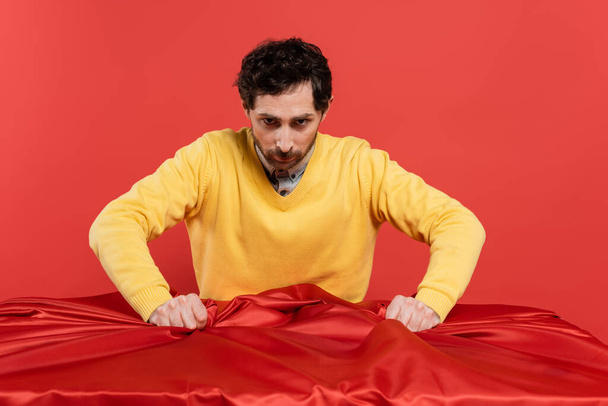 stressed man in yellow jumper pulling red tablecloth on desk isolated on coral background  - Foto, Imagem