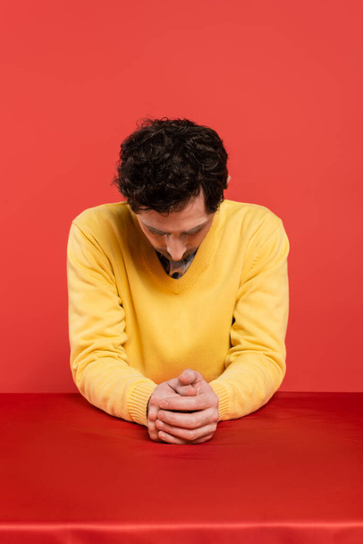 man in yellow long sleeve jumper sitting with bowed head at table isolated on coral background  - Foto, Imagem