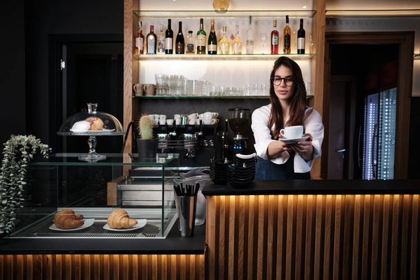 Portrét baristy stojícího u baru v kavárně a držícího šálek čerstvé kávy. - Fotografie, Obrázek