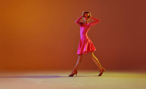 Jive. Estúdio tiro de menina, 11 anos de idade criança em fase festiva vestido dança salão de baile sobre fundo marrom em luz de néon. Conceito de beleza, danças profissionais - Foto, Imagem