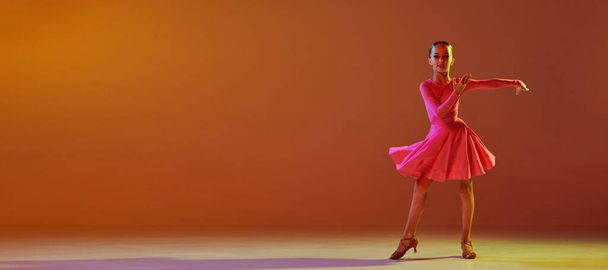 Studioaufnahme eines kleinen Mädchens, 11 Jahre alt, in festlichem Bühnenkleid, das über braunem Hintergrund in Neonlicht einen Standardtanz tanzt. Konzept der Schönheit, professionelle Tänze. Banner mit Copyspace für Werbung - Foto, Bild